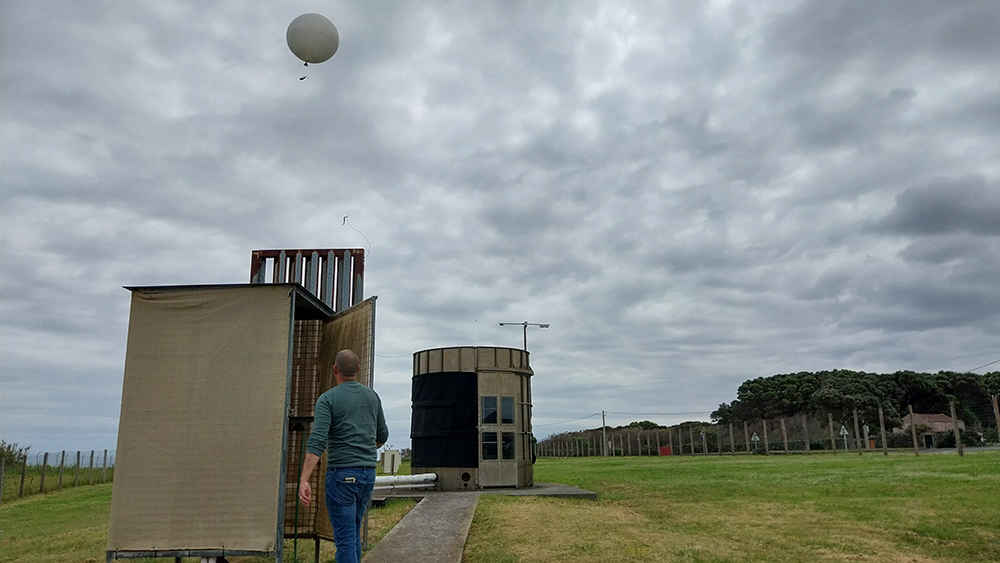 A person looks up at a balloon-borne radiosonde rising into a cloud-filled sky.