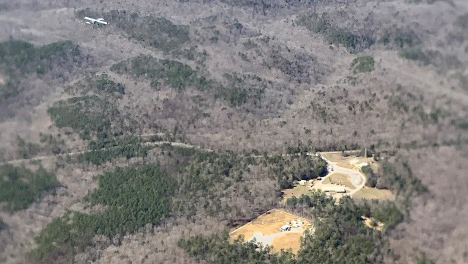 An uncrewed aerial system flies over a forest with buildings and roads visible in the middle of clearings. Patches of brown and green are seen throughout the forest.