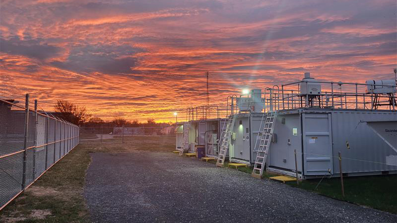 ARM instruments and containers line the edge of grass and gravel path. The sky is orange and cloudy. Street lights are seen in the distance.