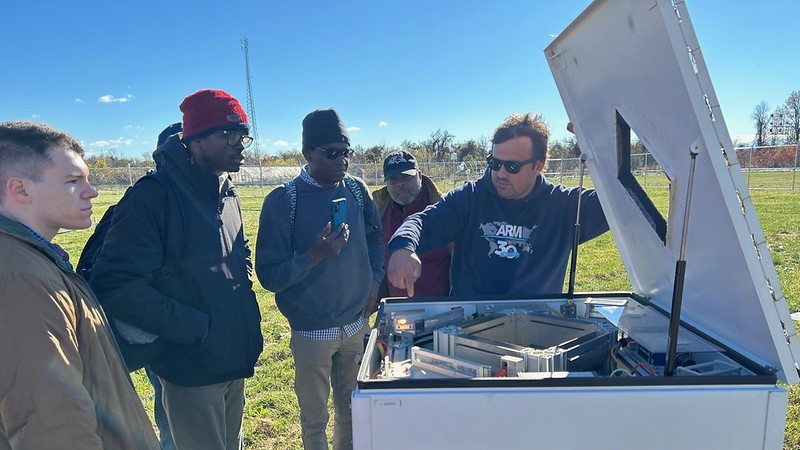 An ARM technician points to a technical component within a video disdrometer while speaking to a group of five people. A green field and fence line sits in the background.