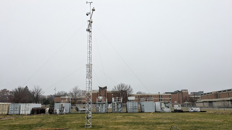 On an overcast day, a tower and large containers outfitted with instrumentation stand on a grass field in front of an academic building.