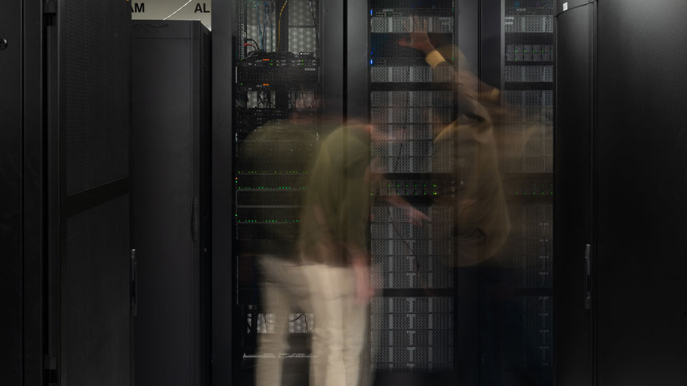 Two people examine a high performance computing cluster. The people are blurry, indicating that they are moving around at a fast pace.