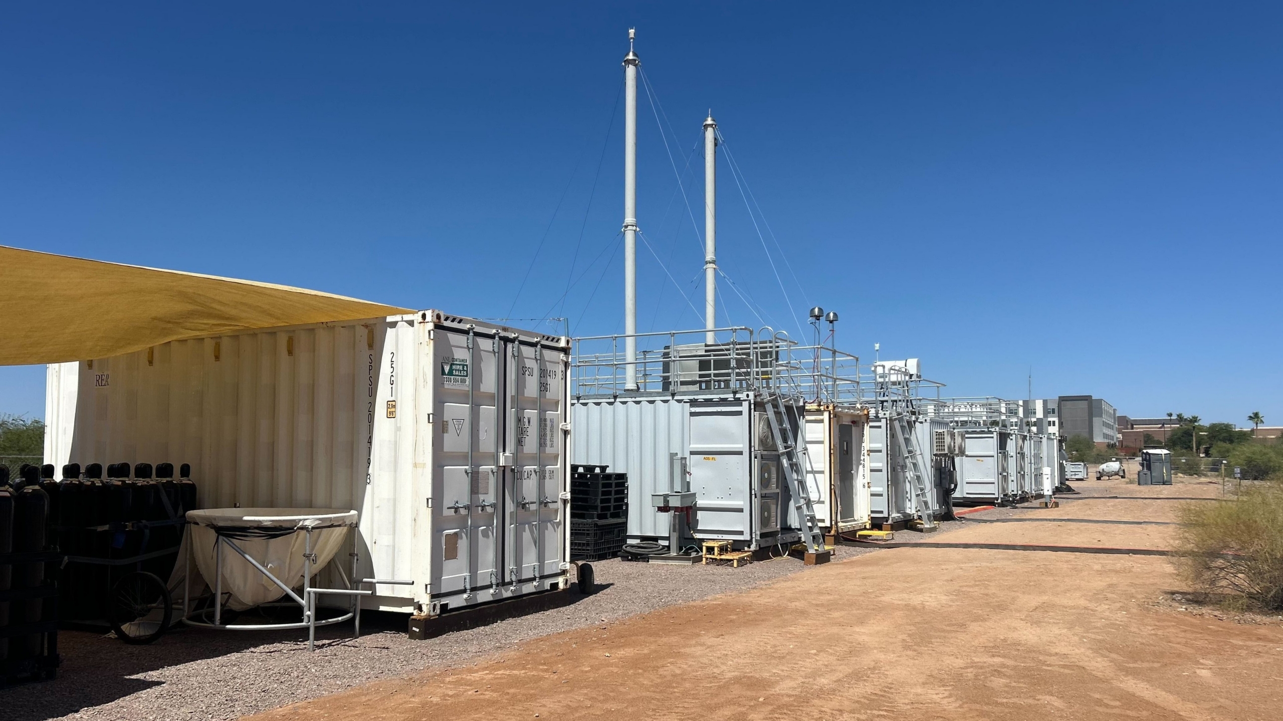 Shipping containers containing atmospheric sensing instruments are installed on a dirt patch with a blue sky above