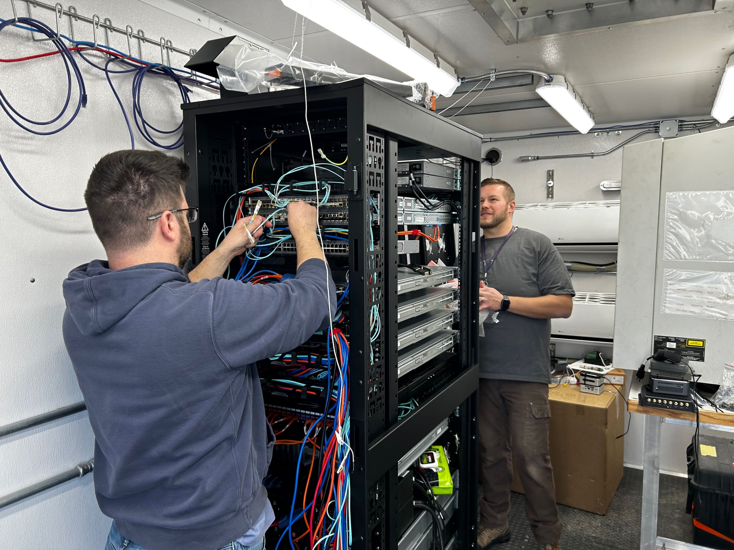 Two people work on a site data system, with multiple cables and wires connected to the system boxes.