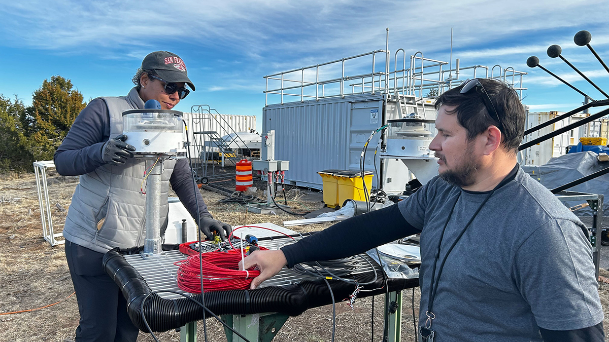 On a sunny day with clouds, one person is standing on a stepstool working on a radiometer, and another is grabbing a red coiled-up cable sitting on the worktable.
