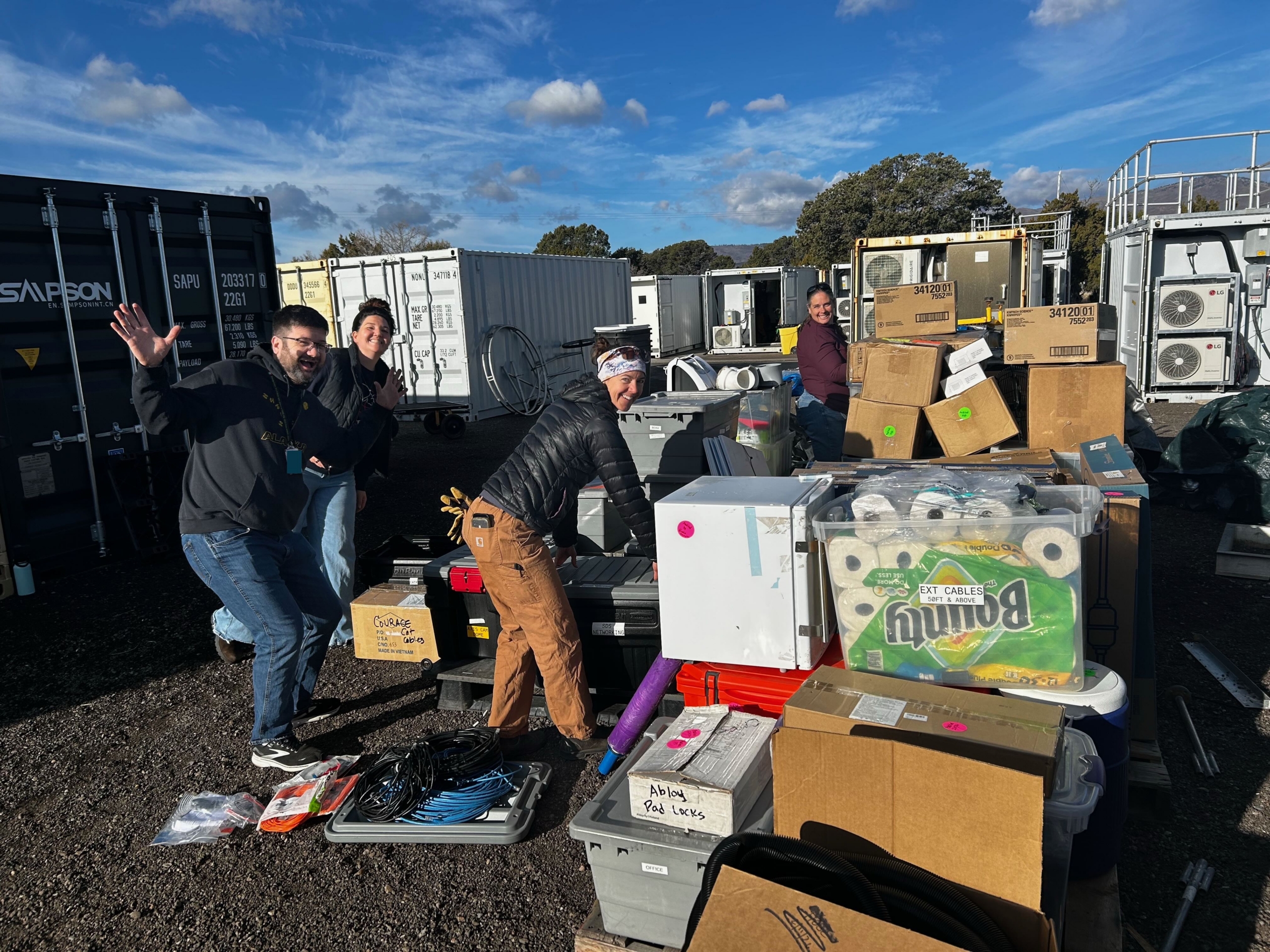 On a sunny day with some clouds in the sky, four people pause to smile at the camera while surrounded by boxes and storage containers.