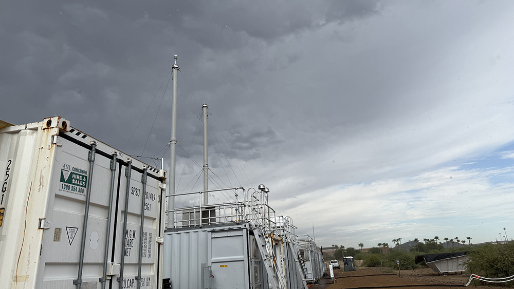Dark clouds sit over containers and atmospheric instrumentation, including a pair of aerosol sampling stacks, with shrubs, palm trees, and a modern academic building nearby.