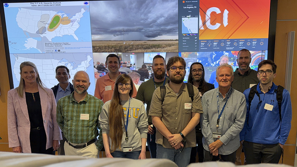 Members of ARM's new data quality operations organizational chart pose for a picture in front of a large screen with six different images showing. Fully visible images include a U.S. hazards outlook map, storm clouds, and icons illustrating weather in different cities.