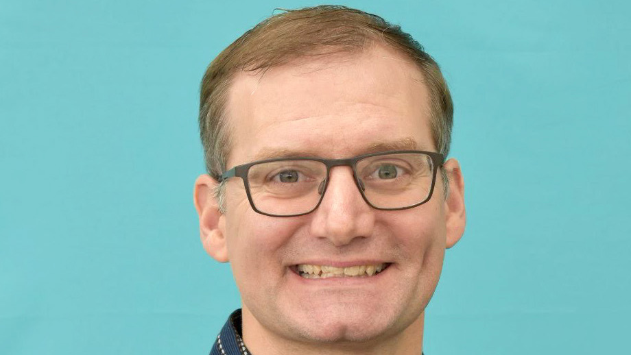 Portrait of Ken Kehoe wearing a striped shirt in front of an aqua background