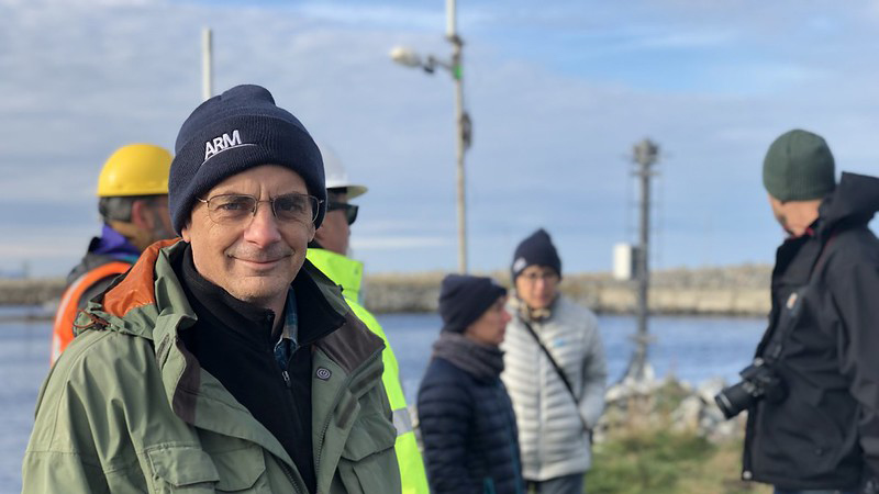 Jim Mather wears an ARM beanie and green jacket and looks at the camera on a cold, cloudy day with others in the background wearing beanies and jackets. A person behind Jim is wearing a safety vest and yellow protective helmet.