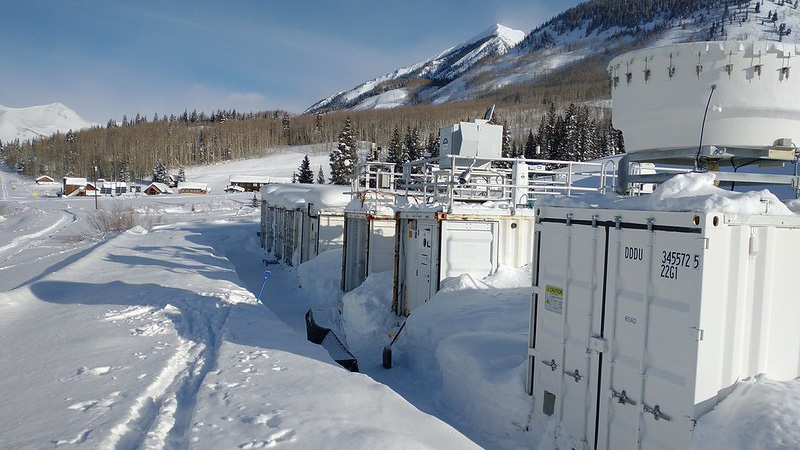 Snow is piled up in between large instrument containers, and ski tracks cut through snow in front of the containers. Trees, buildings, and a snow-covered mountain are visible in the distance on a mostly sunny day.
