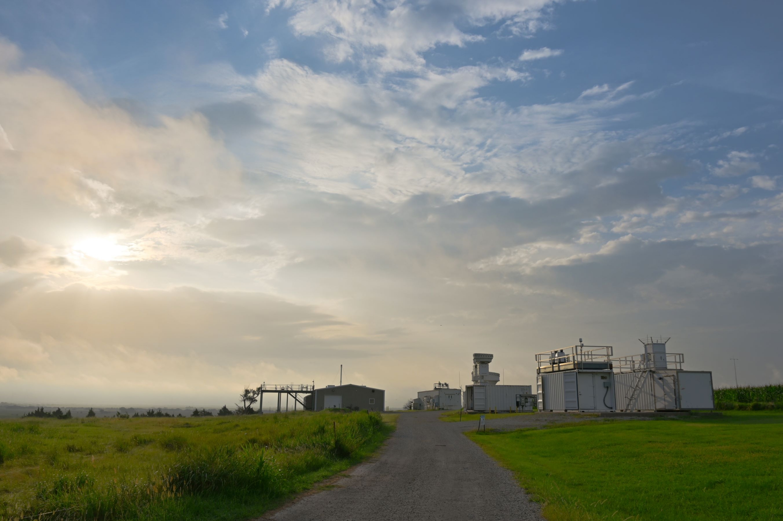A blue sky with clouds and sunlight are pictured above atmospheric instruments and a gravel road