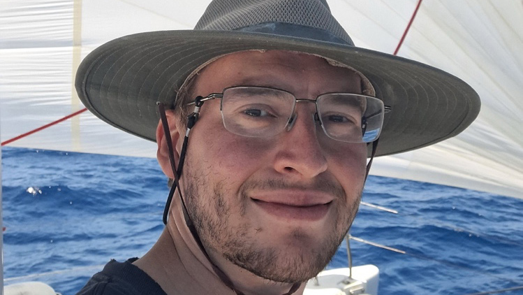Steven Sharpe wears a sun hat, shirt, and glasses on a sailboat with water behind him.