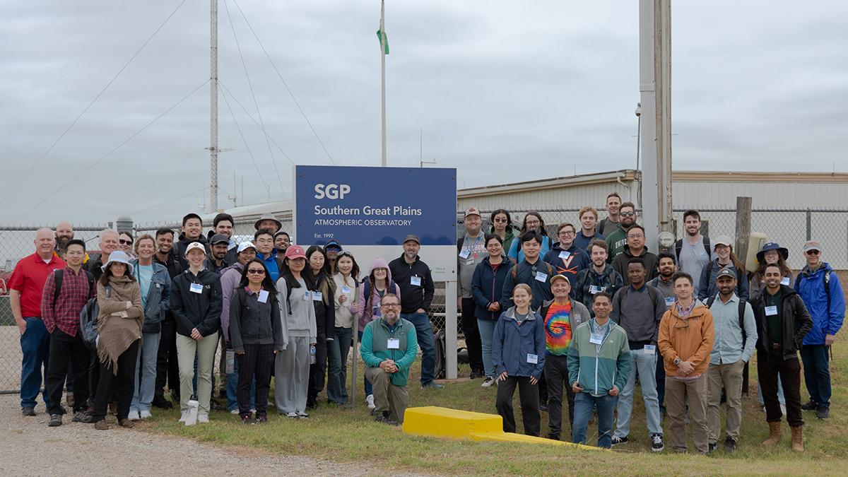On a cloudy day, a group of people stands around a large sign near a gravel path. The sign says, "SGP, Southern Great Plains atmospheric observatory, Est. 1992." A person at the front of the group is kneeling.