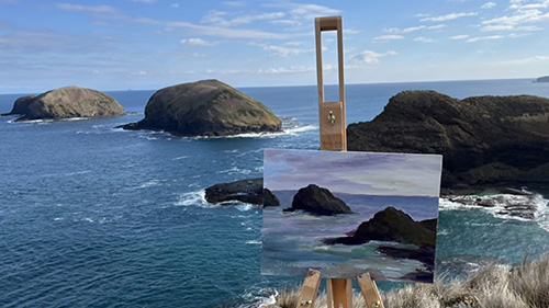 A canvas sits on an easel near a cliff edge. The canvas displays a painted scene that captures the rocks, clouds, and ocean in front of it.