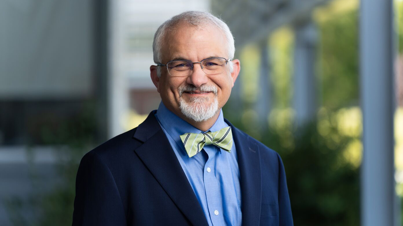 Portrait of Larry Berg wearing a green bowtie with stripes, a blue dress shirt, navy blazer, and glasses
