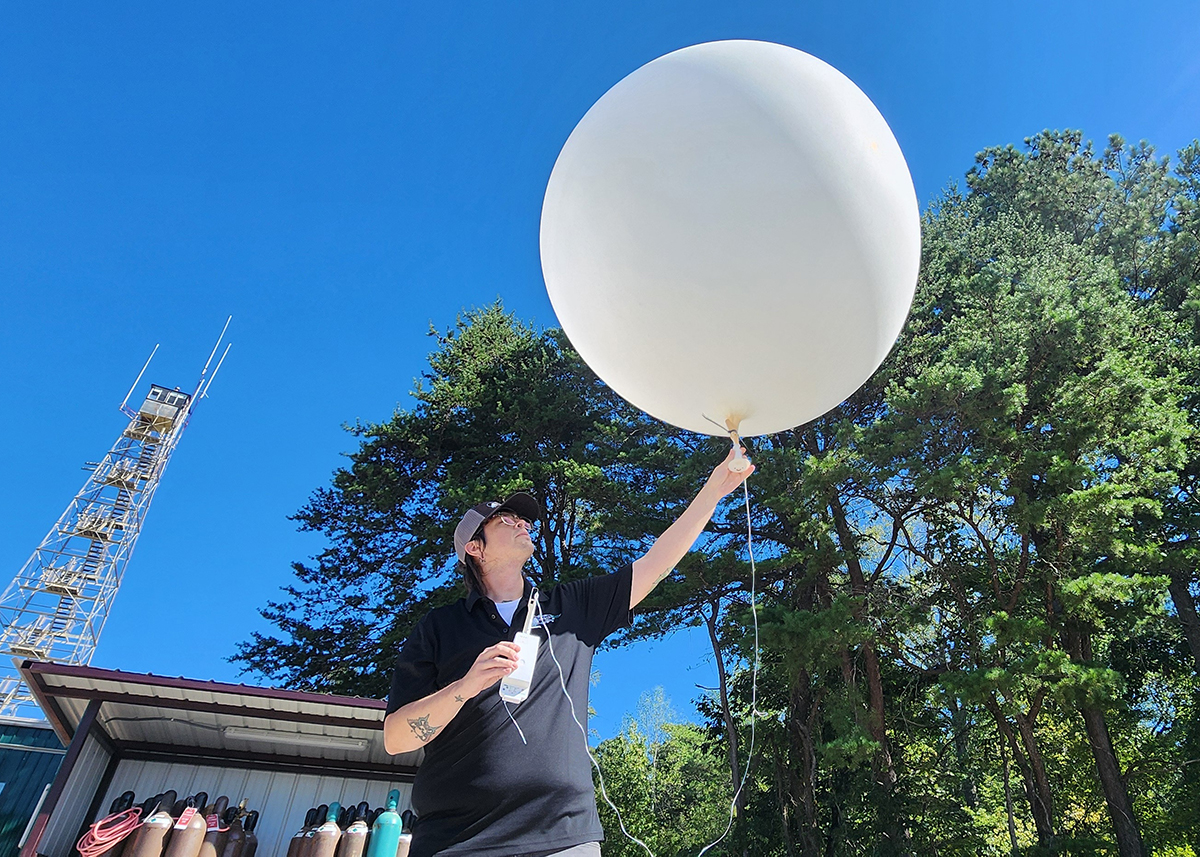 A person holds the bottom of a weather balloon with their left hand and a radiosonde in their right hand. The person, wearing a baseball cap, is looking up at the weather balloon on a sunny day. A tower, helium canisters, and trees are behind the person.