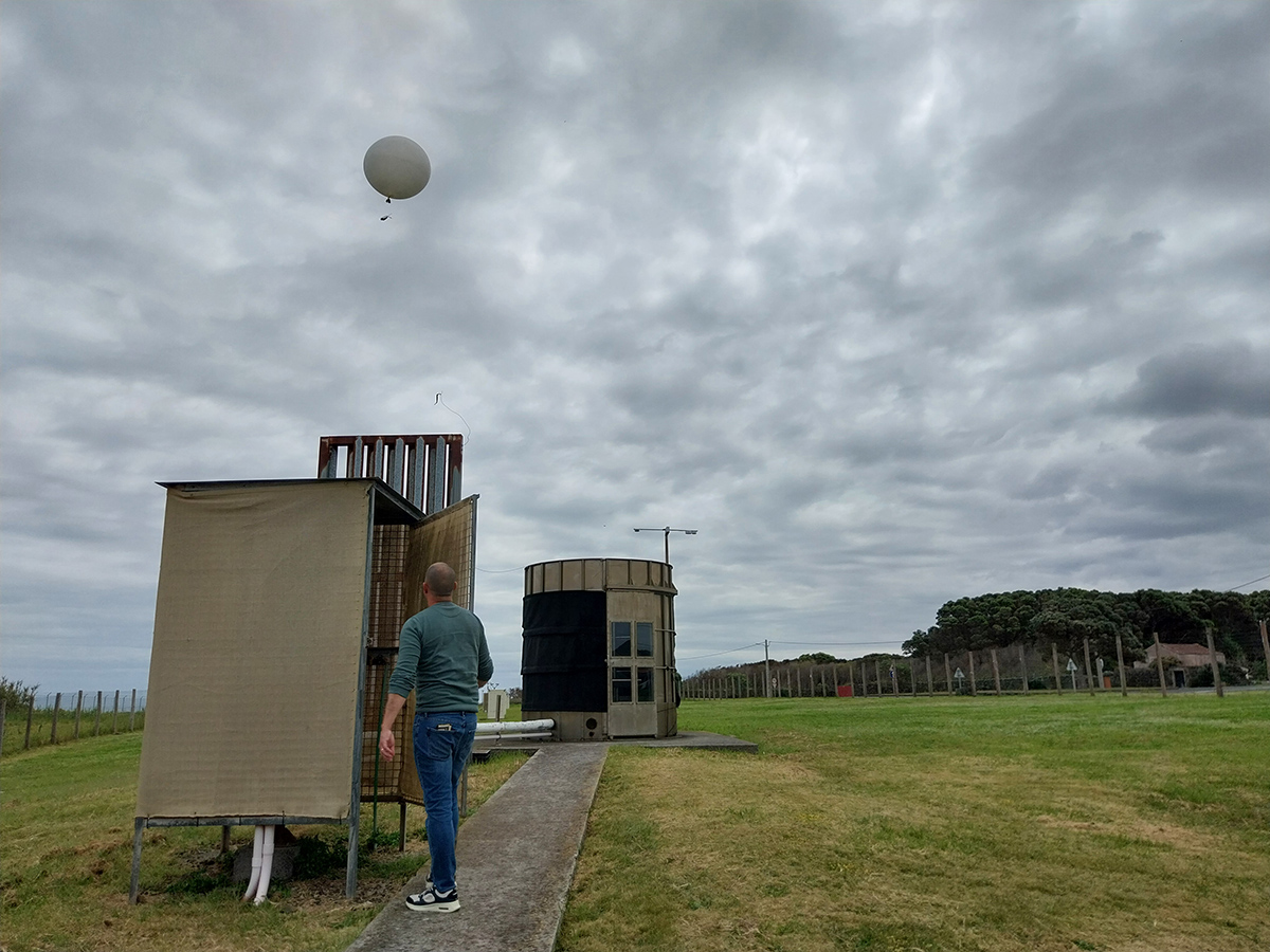 A person looks up at a balloon-borne radiosonde rising into a cloud-filled sky.