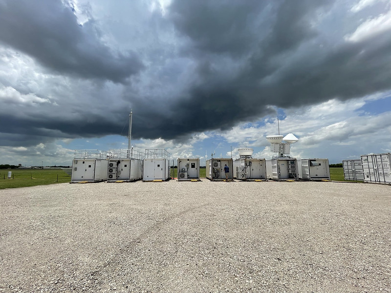 A series of shipping containers outfitted with sophisticated atmospheric and meteorological instruments sits on a gravel clearing with storm clouds overhead.