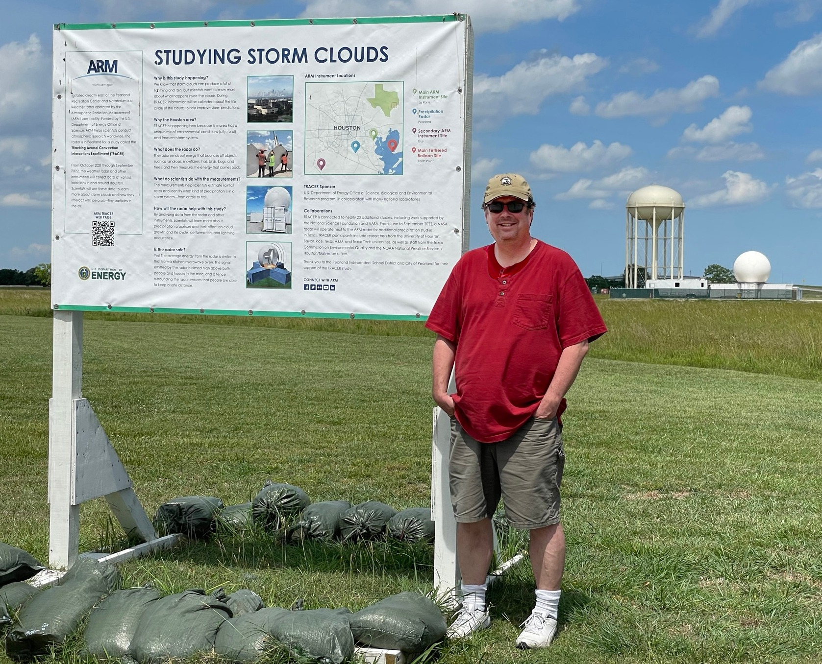 A person stands next to a poster titled, "Studying Storm Clouds." A water tower and a C-band radar are visible in the distance.