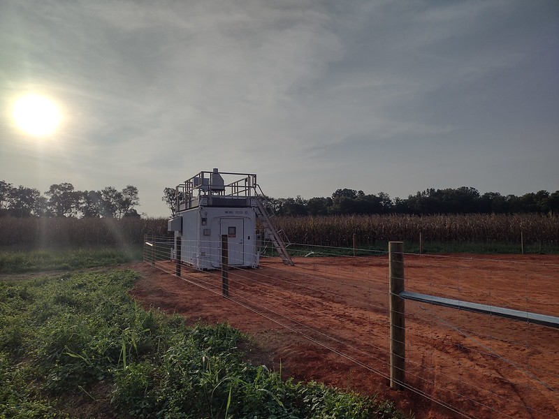 A lidar sits on top on an instrument container sited on dark red soil. Just to the left of the container, the sun streaks through the clouds.