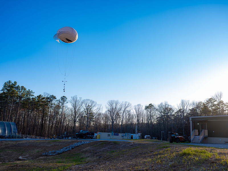 A tethered balloon system hovers over a forest clearing.