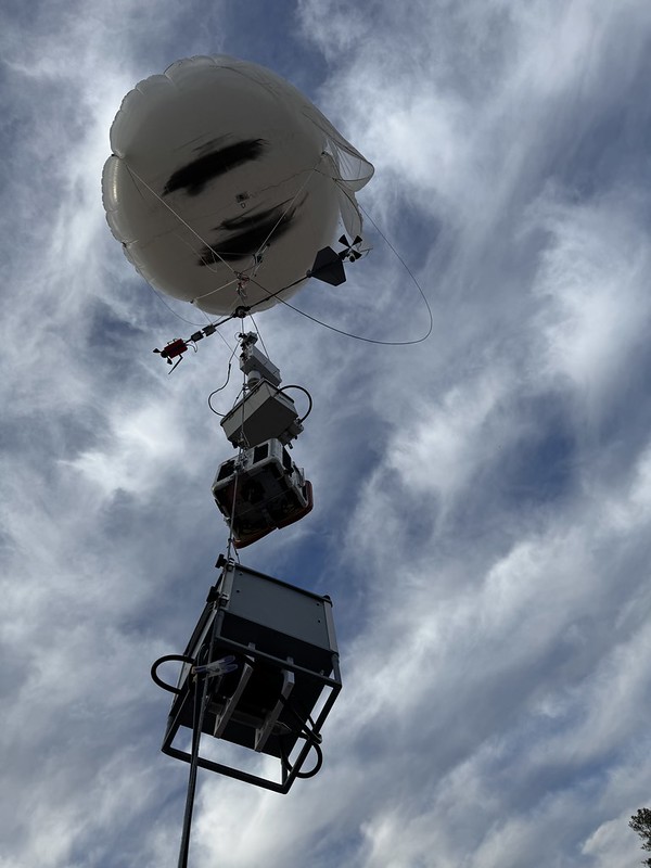 A closeup shot of a balloon with clouds above and instruments strung along the tether below