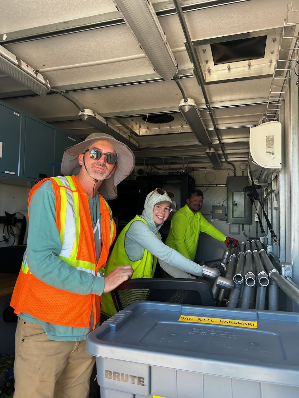 Three people inside a large cargo container stop what they are working on to look at the camera.