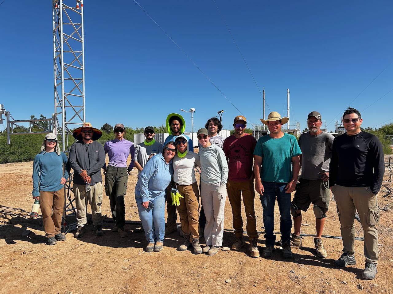A group of people pose in front of atmospheric instrumentation and containers on a cloudless day. They are standing on solid dirt with trees behind them.