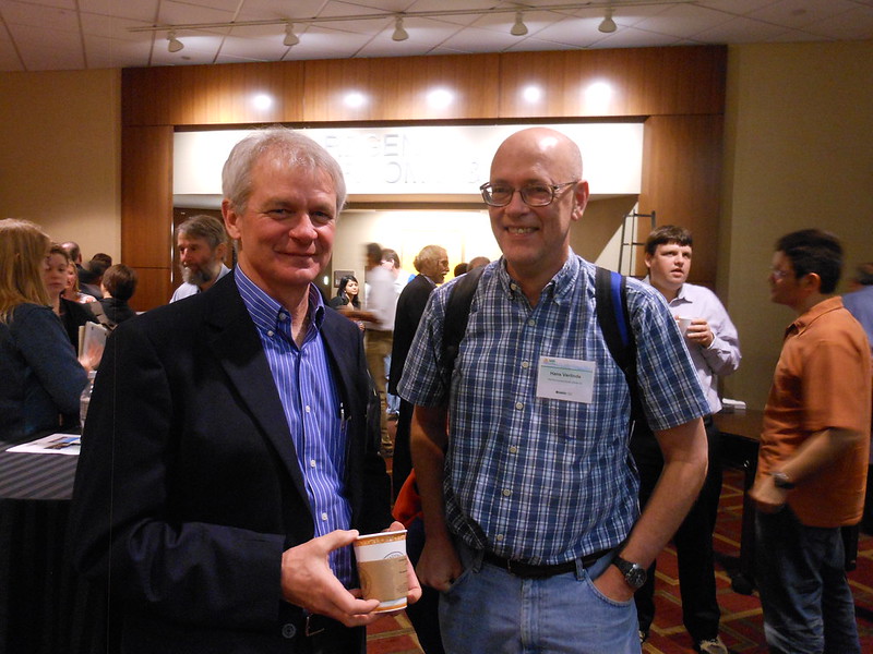 Mark Ivey holds a coffee cup while standing next to Johannes Verlinde in a busy room of people.