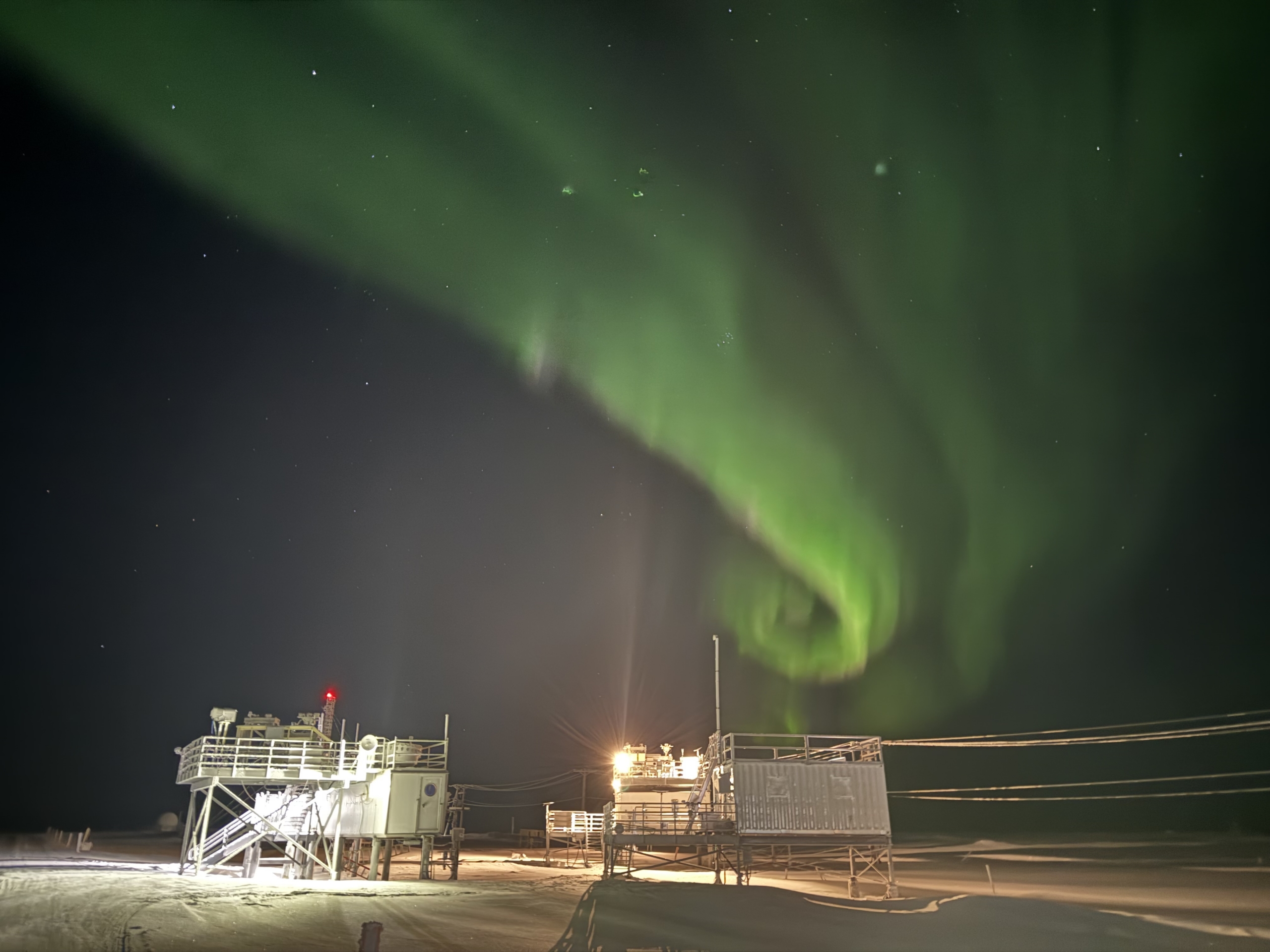 A green aurora streaks, swirls, and glows over instrument platforms on the snow.