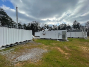 Containers holding atmospheric instruments in a field with green grass. A forest sits in the background.