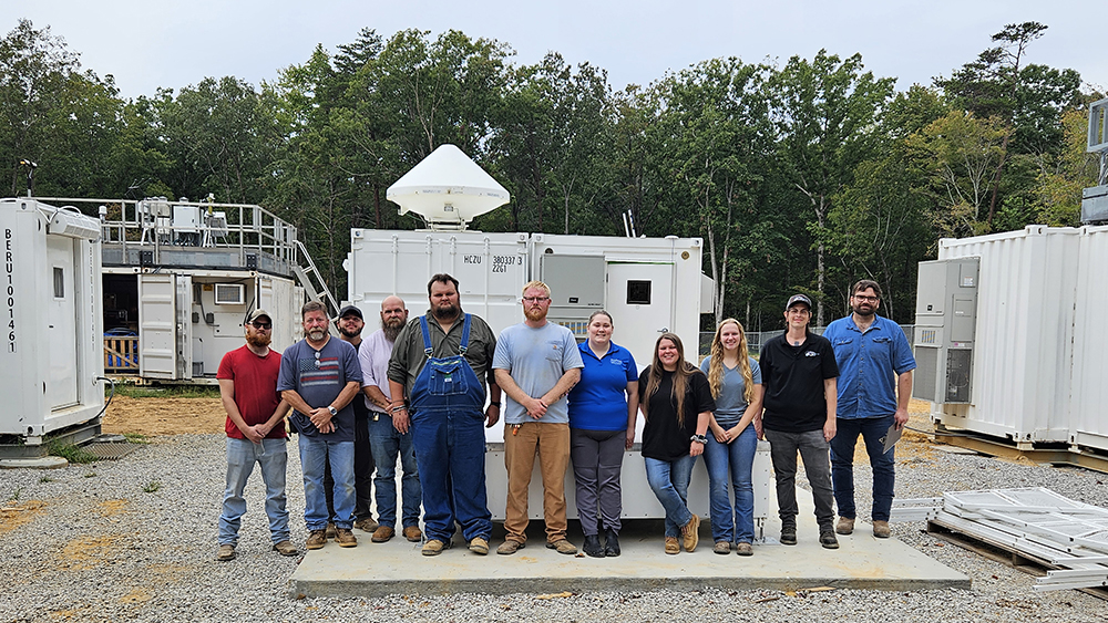 A group of people stand together in front of ARM containers and instruments inside a forest clearing.