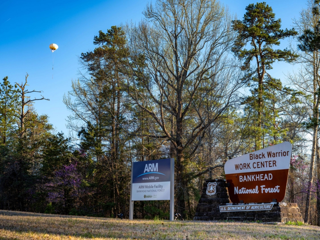 ARM's tethered balloon system flys in a clear sky above trees and signs reading "ARM Mobile Facility Bankhead National Forest" and "Black Warrior Work Center Bankhead National Forest"