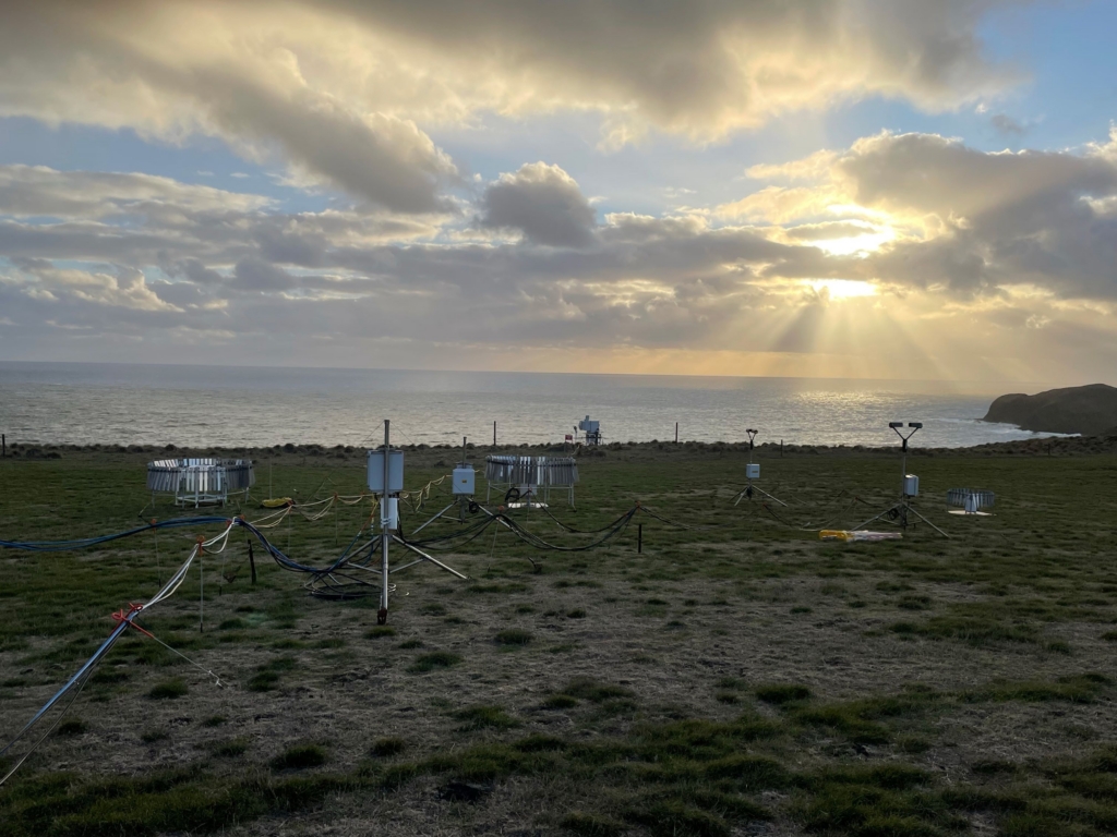 Atmospheric instruments are displayed in a grassy field with an ocean sunset in the background
