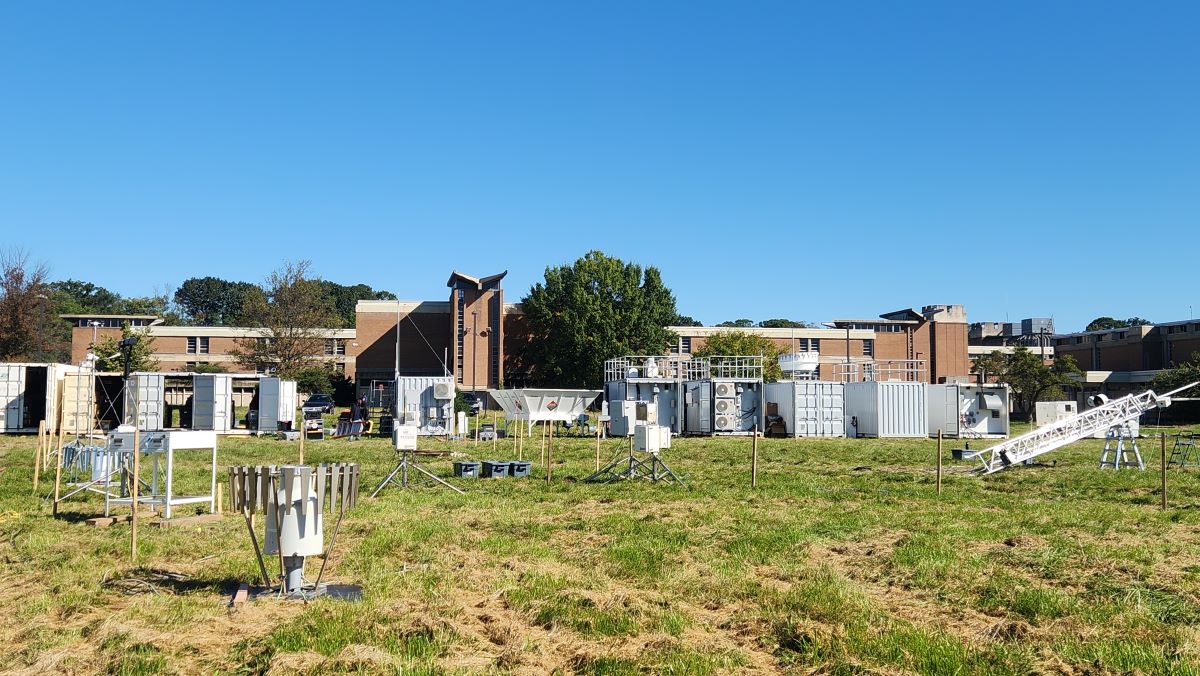 On a sunny day, atmospheric instruments fill a grassy field in front of a line of large instrument containers. An academic building is behind the instrument site.