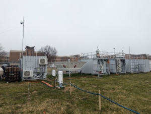 Containers holding atmospheric instruments sit on a grassy field with a brick building in the background