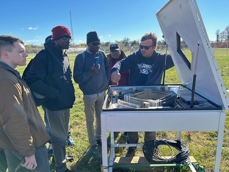 An ARM technician points to a technical component within a video disdrometer while speaking to a group of five people. A green field and fence line sits in the background.