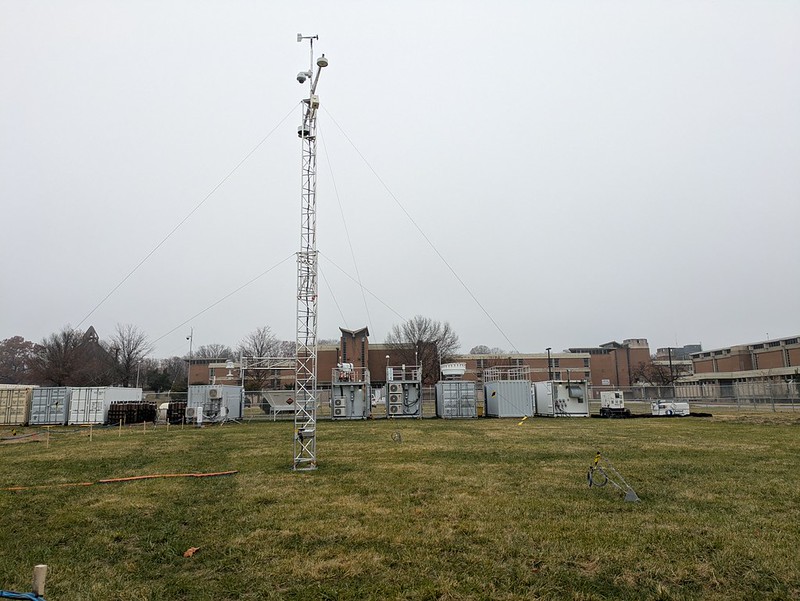On an overcast day, a tower and large containers outfitted with instrumentation stand on a grass field in front of an academic building.