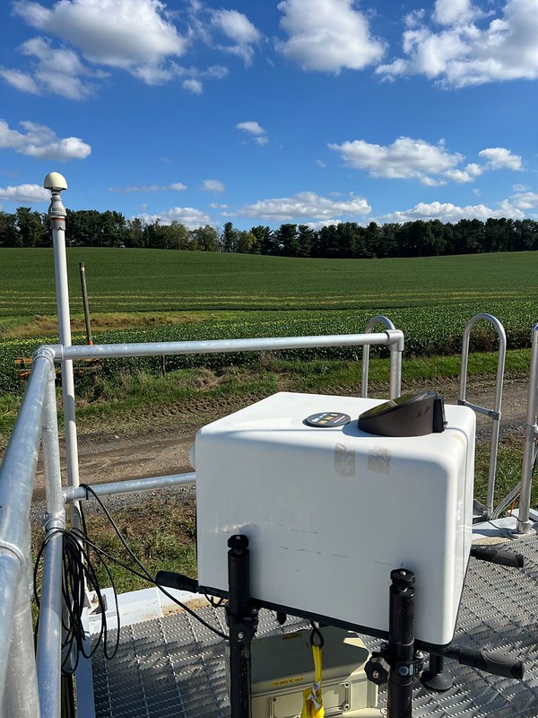 A Doppler lidar sits on an railed instrument platform overlooking farmland with trees along the horizon. Clouds dot the sky overhead.