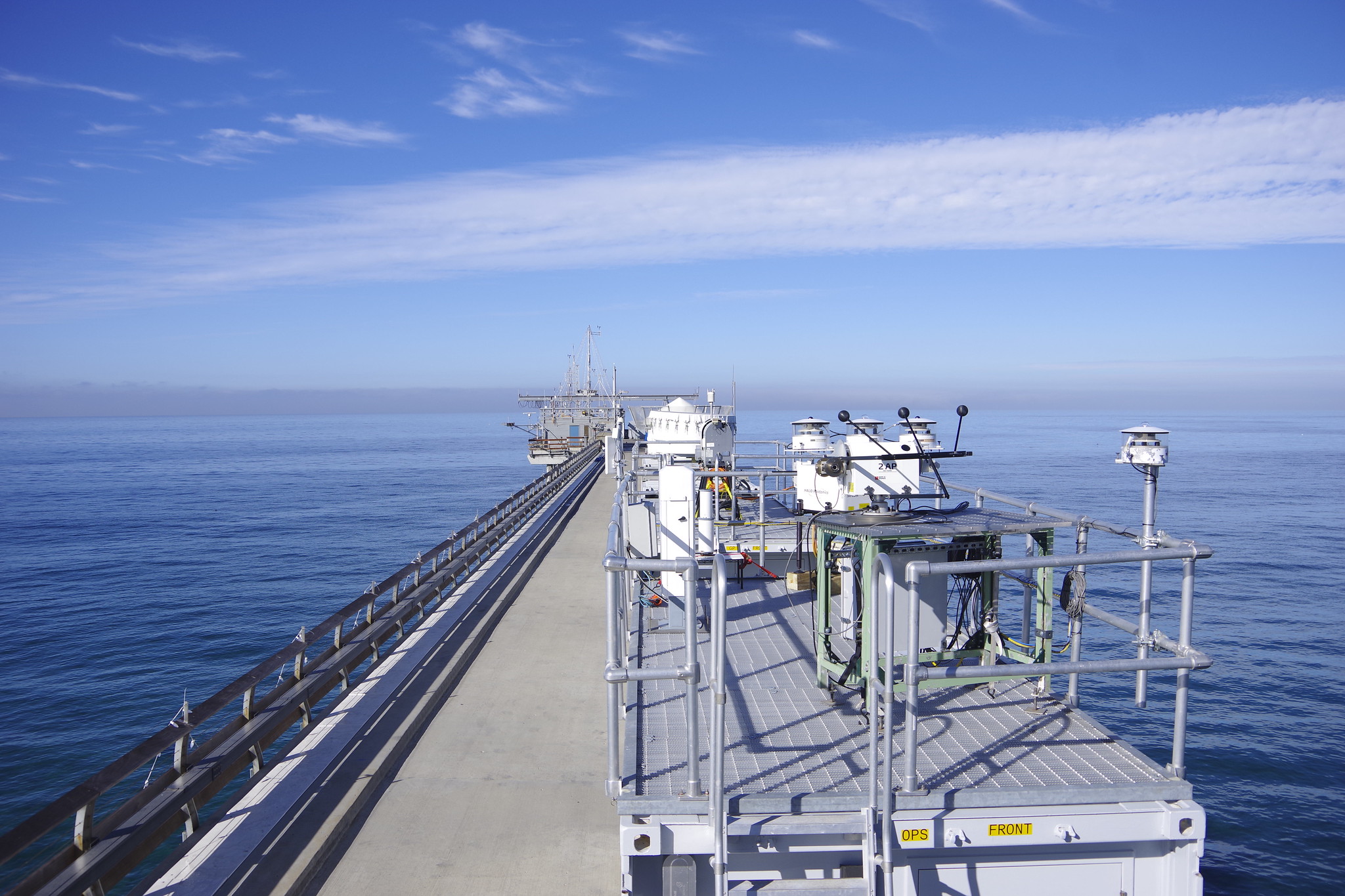 Radiometers and other ARM instruments and containers are situated on a pier surrounded by the Pacific Ocean. A wall of gray clouds is visible along the horizon. A white cloud streaks above the water.