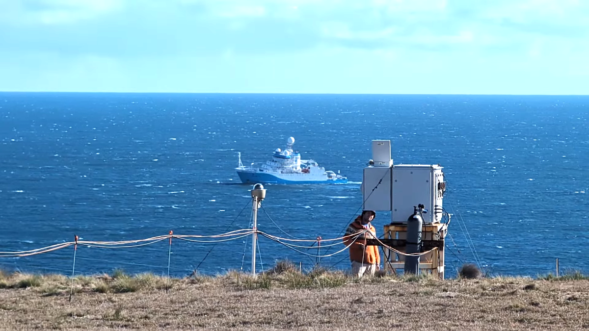 A person stands near a cliff edge looking up at an atmospheric instrument. A large ship is seen on the ocean behind the person.