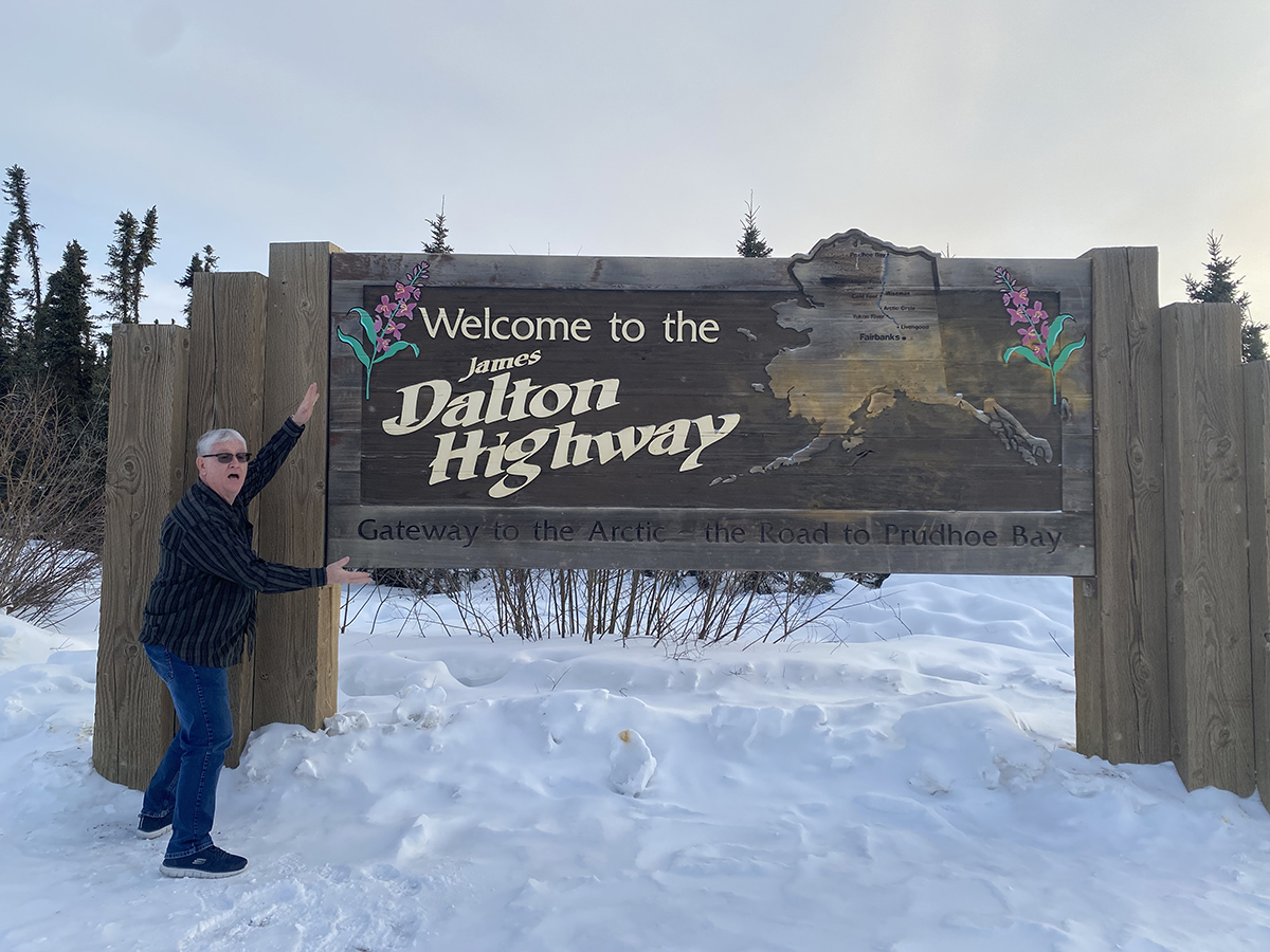 Fred Helsel stands sideways on snow and frames a wooden Dalton Highway sign with his hands.