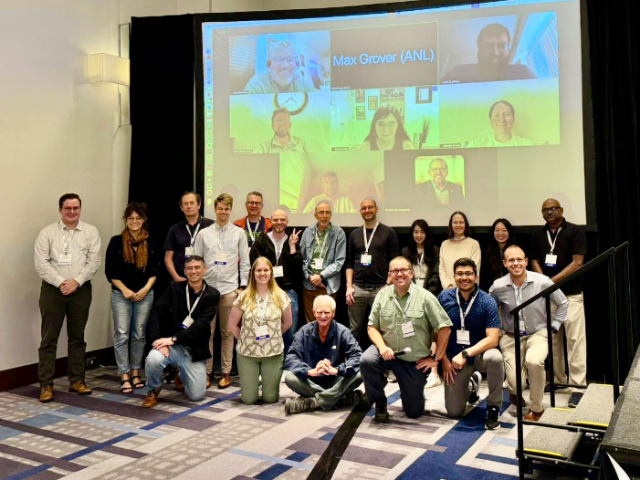 People pose in front of a large screen filled with boxes showing virtual attendees.