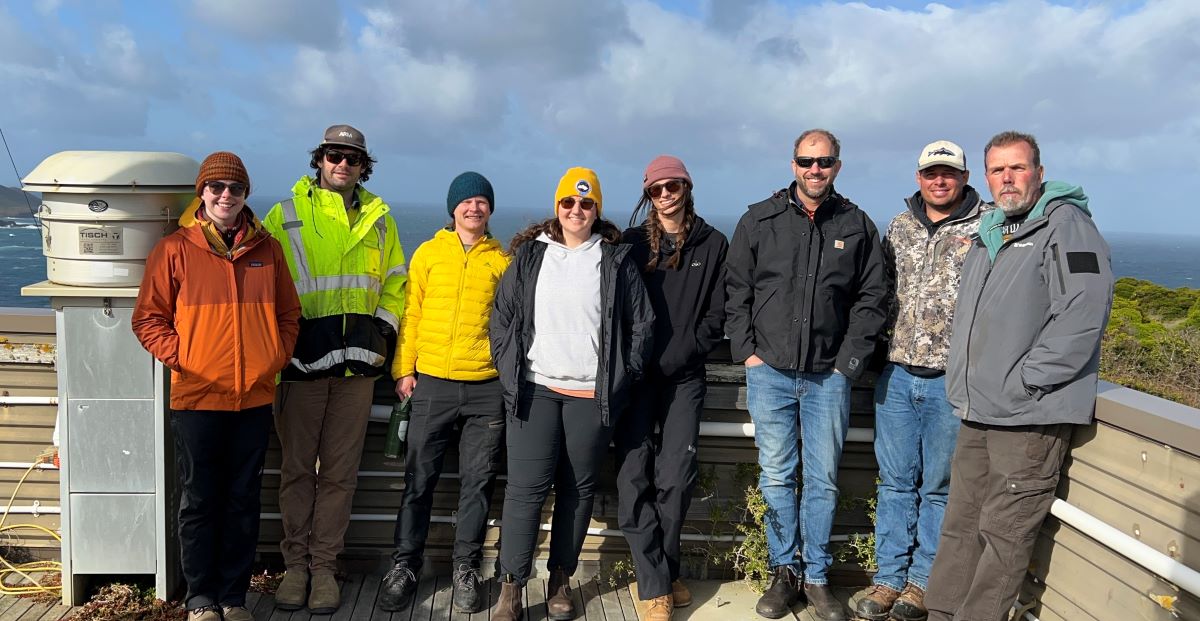 Eight people wearing jackets and pants stand together on a deck with clouds and the ocean behind them.