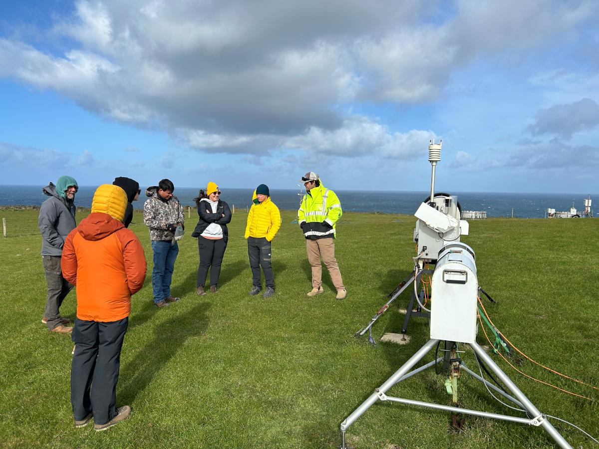 A group of people stands on grass with instruments surrounding them. The ocean is seen behind them.