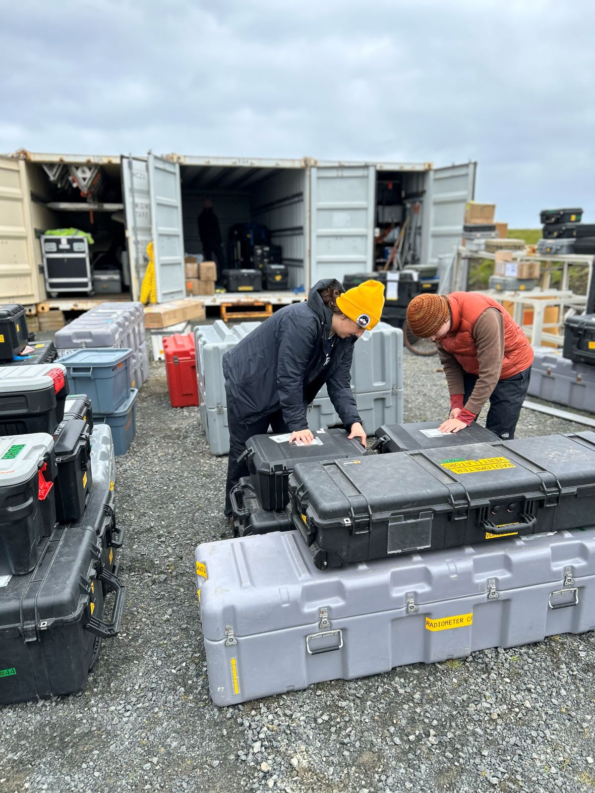 Two people work amid several stacks of instrument cases. Behind them, the doors of large containers are open so the people can load in the instrument cases for shipping.