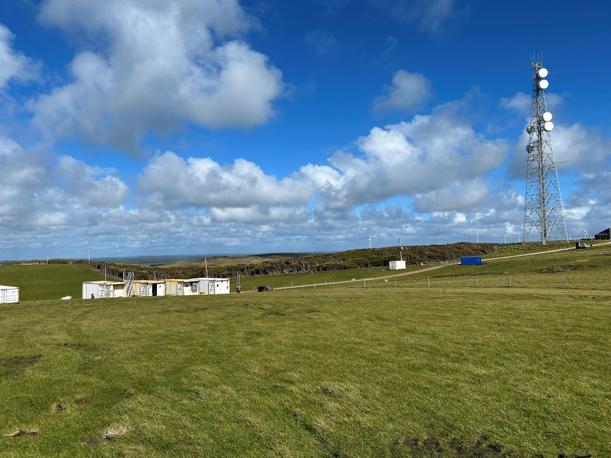 Fluffy clouds float over ARM instrument containers and a large tower on a grassy expanse of land. The ocean peeks out in the distance.