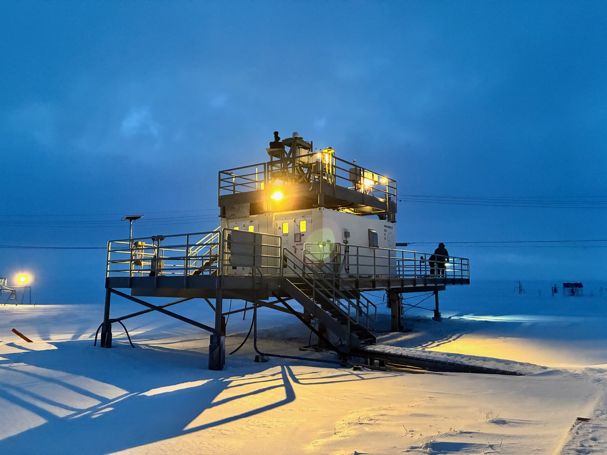 Lights illuminate an instrument deck and the snow below as two shadowy figures work in a corner of the deck.