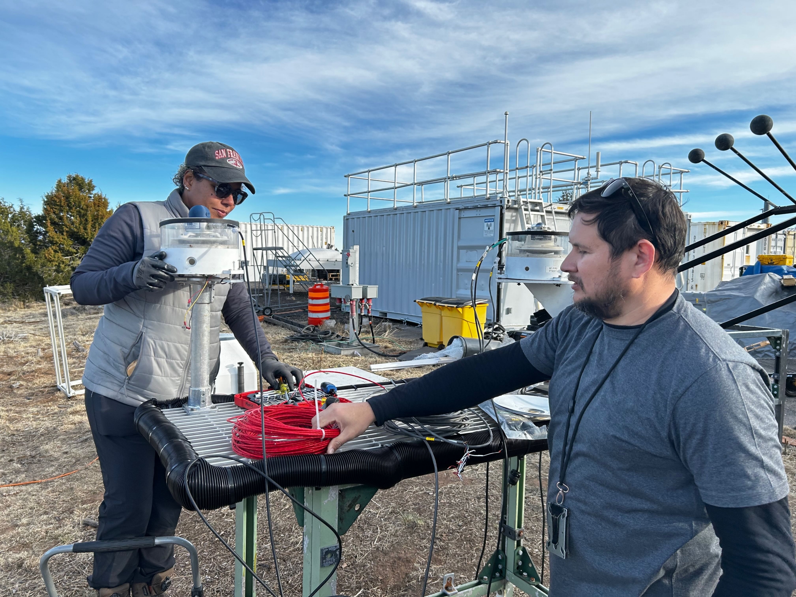 On a sunny day with clouds, one person is standing on a stepstool working on a radiometer, and another is grabbing a red coiled-up cable sitting on the worktable.